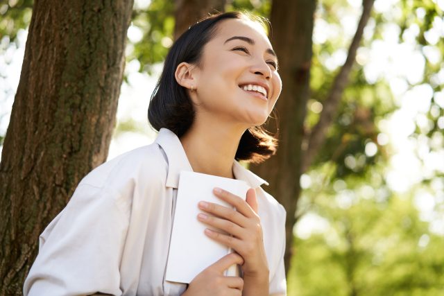 Romantic smiling girl reading book in park or foret, sitting under tree shade on sunny day, relaxing on fresh air surrounded by nature